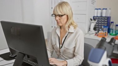 Laboratuvardaki sarışın bir kadın gözlüğünü çıkarıp ekranda gülümsüyor ve klinik ortamdaki iş memnuniyetini yansıtıyor..