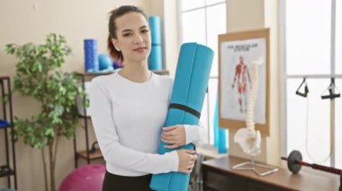 Kendine güvenen genç bir kadın, parlak bir spor salonunda yoga minderi tutuyor ve spor salonuna hazırlanıyor..