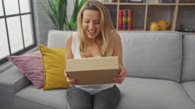 Sarışın bir kadın, elinde karton bir kutu tutarken gülüyor. Gri bir kanepede oturmuş, oturma odasında oturuyordu..