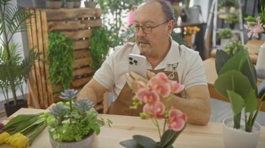 Mature man with glasses using smartphone in a florist shop surrounded by plants clipart