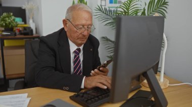 Ofiste çalışan, bilgisayar ve akıllı telefon kullanan, takım elbise ve kravat takmış, etrafı doküman ve bitkilerle çevrili yaşlı, beyaz bir adam görevlerine odaklanmış..