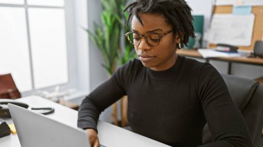 Modern ofis ortamında dizüstü bilgisayar kullanan odaklı Afrikalı Amerikalı kadın.