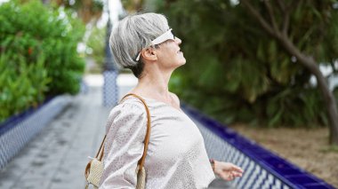 Gri saçlı, zarif, yaşlı İspanyol kadın yemyeşil bir parkta rahatça yürüyor..