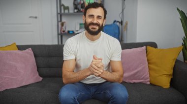 Sakallı, gülen bir adam, gri bir kanepede oturur, samimi ve cana yakın bir tavır sergiler..