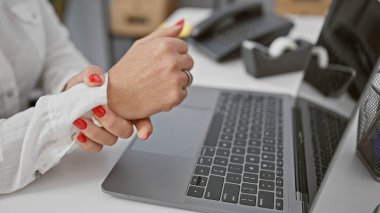 A woman experiencing wrist pain at an office workplace with a laptop, phone, and desk accessories. clipart