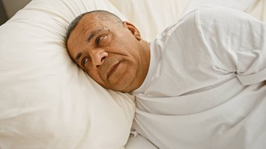 Orta yaşlı İspanyol bir adam beyaz bir yatak odasında dinlenirken dalgın görünür..