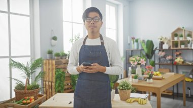 Önlüklü genç bir Asyalı elinde akıllı bir telefonla kapalı bir çiçekçide duruyor..