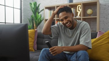 Neşeli bir Afrikalı Amerikalı adam modern bir oturma odasında elinde bir kumandayla gülüyor..