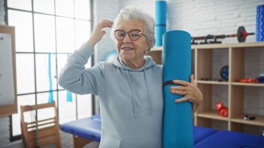 Neşeli, son sınıf öğrencisi bir kadın parlak bir fizyoterapi kliniğinde mavi bir yoga minderi tutuyor..