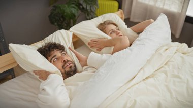 Bir adam ve bir kadın evdeki gürültüyü resmeden yatak odasındaki yastıklarla kulaklarını kapatıyorlar..