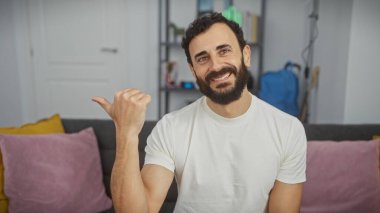 Beyaz tişörtlü, gülümseyen sakallı adam evin içinde, rahat oturma odası arka planında başparmak kaldırıyor.