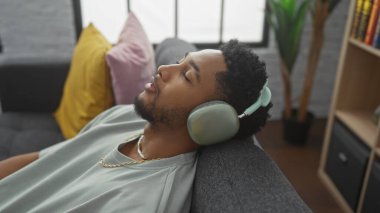 Rahat bir Afrikalı Amerikalı adam rahat bir oturma odasında kulaklıkla müzik dinlemekten hoşlanır..