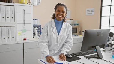 Bir laboratuarda bilgisayar, dosyalar ve laboratuvar ekipmanlarıyla gülümseyen Afrikalı Amerikalı kadın bilim adamı..