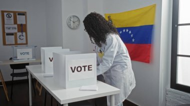 Kıvırcık saçlı Afro-Amerikalı genç bir kadın, Venezuela bayrağı ve pankartı olan kapalı bir seçim odasında oy veriyor..