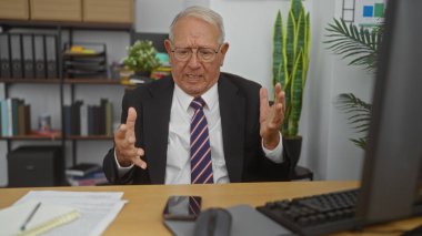 Takım elbiseli ve gözlüklü yaşlı beyaz adam modern ofisinde bir video görüşmesi sırasında hayal kırıklığı içinde el kol hareketi yapıyor..