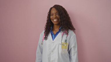 Kıvırcık saçlı, güzel, genç, Afro-Amerikan bir kadın doktor pembe bir arka plana gülümsüyor..