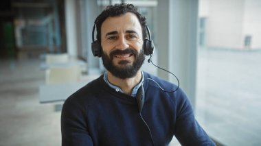 Modern ofis ortamında kulaklıklı, gülümseyen sakallı adam.