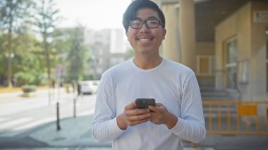 Güneşli bir şehir sokağında akıllı telefon kullanan gülümseyen Asyalı genç adam..