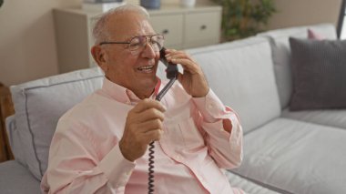 Oturma odasındaki kanepede otururken telefonda gülen yaşlı gri saçlı beyaz adam..