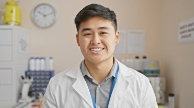 Laboratuvar ortamında genç bir Asyalı bilim adamı kameraya gülümsüyor, laboratuvar önlüğü ve iple sarılı..