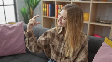 Gülümseyen beyaz bir kadın, küçük boyutta bir ev içi jesti yapıyor, rahat bir ev ortamı sergiliyor..