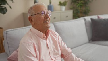Pembe gömlekli yaşlı beyaz adam, gri koltukları ve kapalı bitkileri olan rahat bir oturma odasında oturuyor, pencereye doğru bakarken keyifle gülümsüyor..