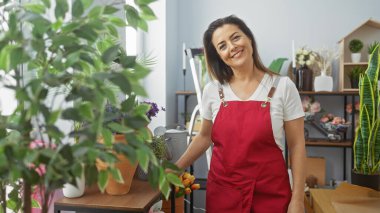 Kırmızı önlüklü, gülümseyen İspanyol kadın ev bitkileriyle çevrili laleler düzenliyor.