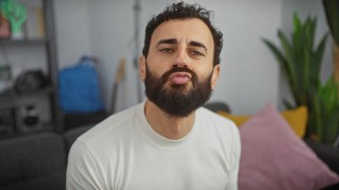 Orta yaşlı, yakışıklı, sakallı bir adam modern bir oturma odasında komik bir surat yapıyor..