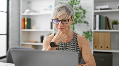 Gri saçlı, gözlüklü, modern ofis ortamında çalışan olgun İspanyol bir kadın..
