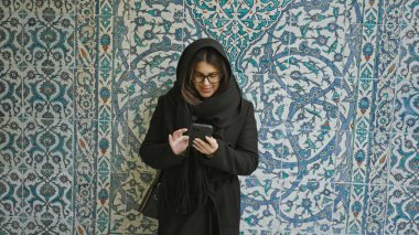 Siyah fularlı genç bir kadın geleneksel Türk fayanslarına karşı akıllı telefon kullanıyor..