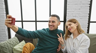 Gülümseyen bir çift, modern bir dairede rahat bir kanepede birlikte selfie çekerler. Aşkı ve birlikteliği tasvir ederler..