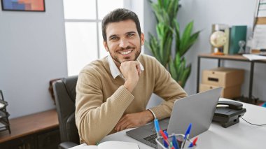 Sakallı, gülümseyen İspanyol bir adam modern ofis ortamında kendine güveni ve profesyonelliği yayarak poz veriyor..