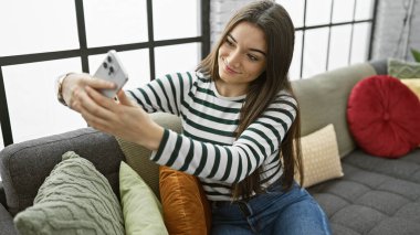 Telefonuyla selfie çekerken gülümseyen genç bir kadın. Kapalı bir kanepede rahatça oturuyor..