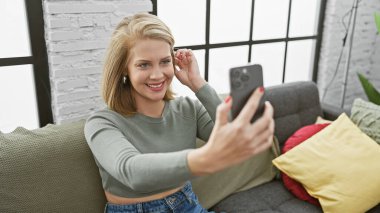 Güzel, genç, sarışın bir kadın akıllı telefonuyla kanepenin içinde selfie çekiyor.