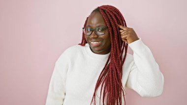 Saçları örgülü neşeli siyah bir kadın kafasına dokunuyor, pembe bir arka plana karşı duruyor gözlük takıyor ve beyaz bir süveter giyiyor..