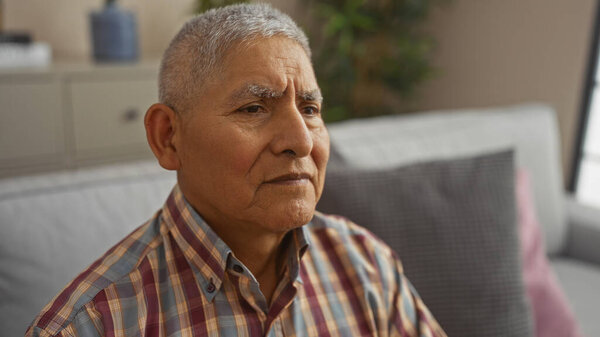 A mature hispanic man with grey hair and a plaid shirt sits thoughtfully in a cozy living room with a comfortable ambiance.