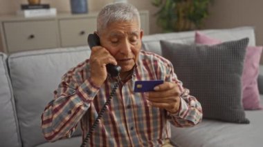 Orta yaşlı İspanyol bir adam rahat bir oturma odasında kanepede otururken elinde kredi kartı ve telefon kullanıyor..