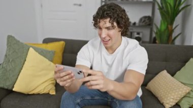 Kıvırcık saçlı İspanyol adam gülümsüyor ve oturma odasındaki kanepede akıllı telefon kullanıyor.