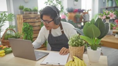 Önlüklü Asyalı bir adam çiçekçide notlar alıyor bitkiler, dizüstü bilgisayarlar ve kulaklıklarla..