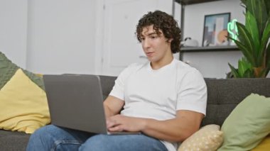 Kanepede oturan Latin asıllı genç adam dizüstü bilgisayar kullanıyor. Korkmuş ve ağzı açık bir şekilde hayrete düşmüş. Şaşırmak için.