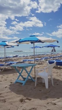 Açık gökyüzü ve deniz manzaralı kumlu bir sahilde mavi şemsiyelerin altında boş güneşlikler..