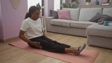 Genç Afro-Amerikalı kadın, bir evin rahat oturma odasında minder üzerinde yoga egzersizleri yapıyor. Günlük siyah beyaz giysiler giyiyor..
