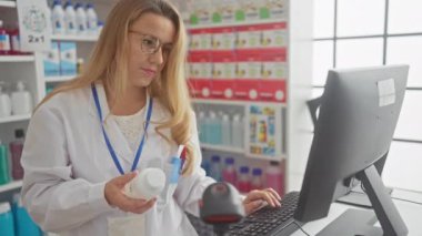 Odaklanmış sarışın bir eczacı modern bir eczanede bilgisayarla çalışırken ilaç şişesini kontrol ediyor..