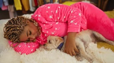 Pembe pijamalı, köpeğiyle rahat bir yatak odasında yatağa uzanmış Afro-Amerikalı genç bir kadın..