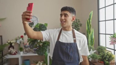 Genç bir adam çiçekçi dükkanında akıllı telefonuyla selfie çekiyor. Etrafı bitkiler ve çiçeklerle çevrili..