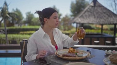 Genç bir İspanyol kadın, Bali tatil köyünün terasında, sakin bir ifadeyle içkisini yudumlarken, sıradan bir açık hava yemek deneyiminden zevk alıyor..