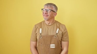 Gözlüklü neşeli gri saçlı adam, düşüncelere dalmış, önlük takmış, ayakta durmuş gülümsüyor, izole edilmiş sarı bir arka plan üzerinden gözlerini kaçırıyor.