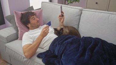 Kedisi yanında rahat bir oturma odasında yatarken, kredi kartı ve telefonuyla koltukta dinlenen bir adam..