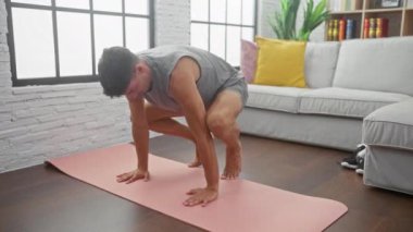 Formda bir adam, parlak, modern bir oturma odasında, rahat bir kanepesi olan pembe bir paspasın üzerinde yoga yapıyor..