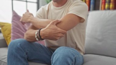 Genç bir İspanyol adam, parlak, rahat bir oturma odasında kanepede otururken kolunu tutarak acı içinde ürker..